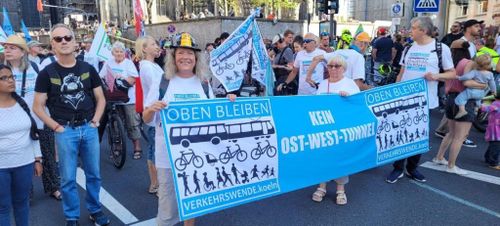 Vorderseite einer Demo des Bündnis Verkehrswende Köln gegen den Tunnelbau auf der Ost-West-Achse auf der Trankgasse zu Fuße des Kölner Doms.

Im Vordergrund halten drei Teilnehmer ein großes hellblaues Banner mit dem Titel "Kein Ost-West-Tunnel". Links und rechts neben dem Titel befindet sich das Logo des Bündnis: Im Kopfbereich steht weiss auf blau "OBEN BLEIBEN" in der Mitte sind schwarz auf weiß Piktogramme von (oben nach unten) Bus, Tram, Fahrrädern (eins mit Anhänger), sowie diverse Fußgänger*innen jeden Alters (teils im Rollstuhl oder mit Kinderwagen) zu sehen.
Im Fußbereich steht weiß auf blau: "VERKEHRSWENDE.koeln".

Einige Teilnehmer schwenken Flaggen mit dem Logo oder tragen es auf weissen T-Shirts.