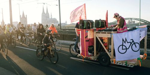 Der Fahrrad-Rave fährt über die Deutzer Brücke richtung Deutz.
Die Spitze der Demo mit dem, mit Flaggen des Fahrrad-Entscheids geschmückten, DJ-Bike und den folgenden Teilnehmern auf ihren Rädern fahren im Vordergrund.
Im Hintergrund ist Groß St. Martin, der Dom und die Hohenzollernbrücke zu sehen.